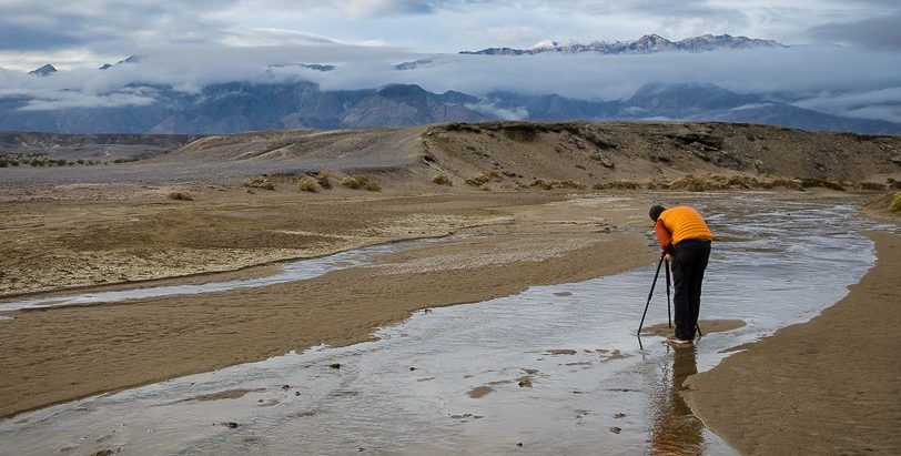 a photographer in a yellow vest stands behind his tripod near Salt Creek in Death Valley National Park. Clouds surround the mountains in the background and the scene is cloudy, dark, and rainy.